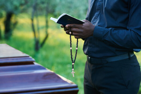 Close-up of African American man in priest attire holding rosary beads and open Holy Bible while standing in front of coffin during burial service