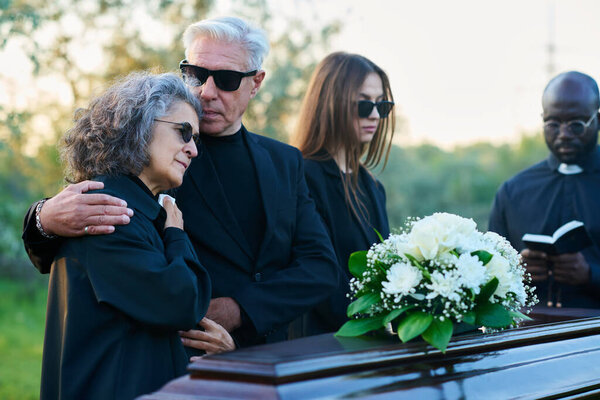 Mourning family of three and priest in black attire standing in front of coffin with white flowers on top during funeral service at graveyard