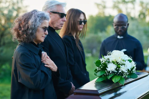 Mourning mature woman with handkerchief keeping her hands on chest while standing in front of coffin with closed lid during funeral service