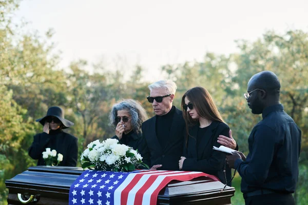 Pastor with Holy Bible and mourning family of three standing in front of coffincovered with USA flag during funeral service at graveyard