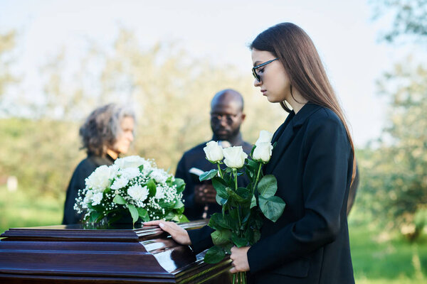 Side view of young mourning woman with bunch of white roses standing by coffin with fresh chrysanthemums on top of closed lid at funeral
