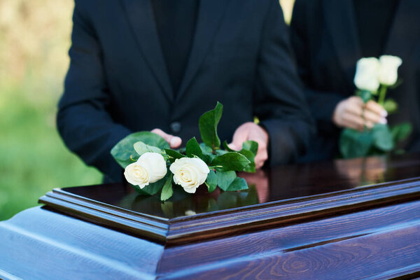 Focus on two fresh white roses being put on coffin lid by mourning mature man wearing black suit standing against young woman with flowers