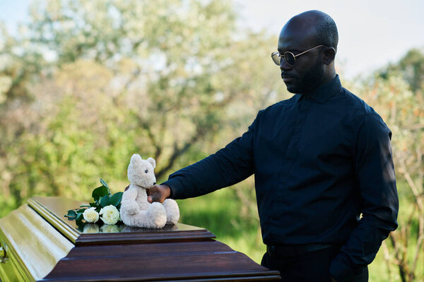 Young grieving African American man in mourning clothes putting bunch of white roses and teddybear on top of closed lid of coffin at funeral