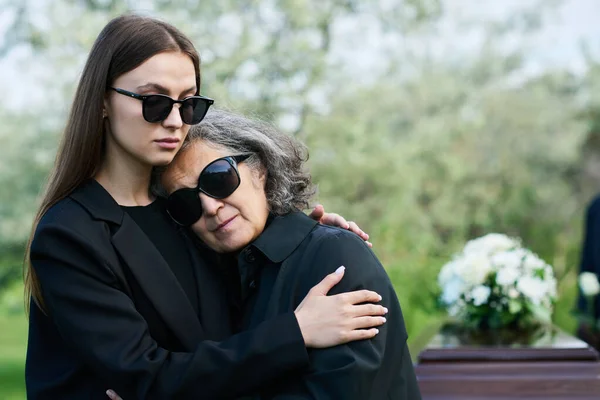 Young woman in sunglasses and mourning attire supporting her grieving mother or grandmother at funeral of their family member or friend