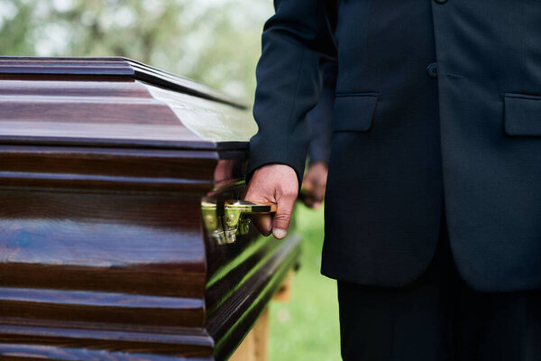 Close-up of man in black suit holding by handle of wooden coffin with dead person while carrying it with other people at funeral ceremony