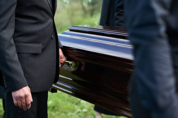Focus on man in black suit holding by handle of wooden coffin while carrying it together with other people after farewell ceremony
