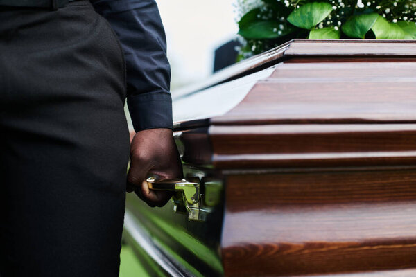Close-up of man holding wooden coffin by one of several handles while carrying it with other people after farewell ceremony at funeral