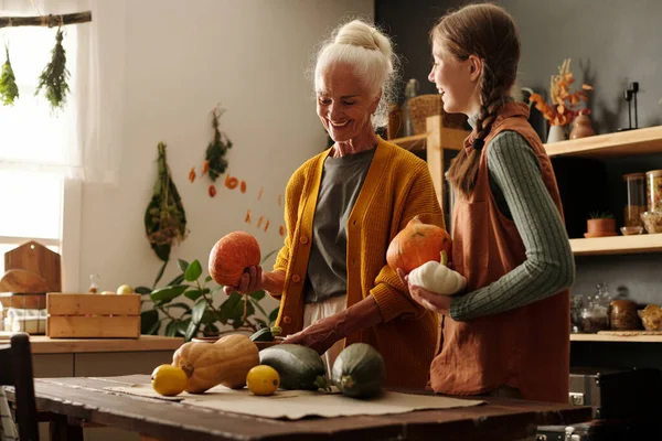 stock image Happy aged woman and her cute granddaughter with pigtails having chat while standing by kitchen table and sorting out fresh vegetables