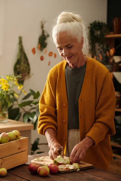 stock image Senior woman with white hair standing by wooden table in front of camera and cutting fresh ripe apples grown in the garden by country house