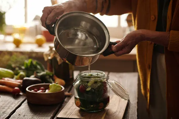Hands of senior woman with metallic pan pouring hot water into jar with fresh garden herbs and vegetables while preparing pickles for winter