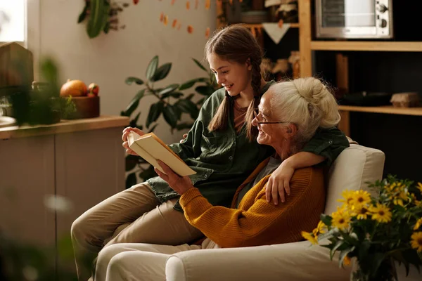 坐在舒适的扶手椅上悠闲地阅读故事书或小说的快乐祖母和孙女的侧视图 — 图库照片