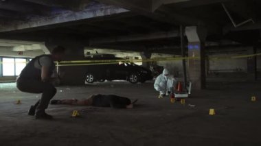 Tam bir erkek polis dedektifi cinayet mahallinde dolanıyor, yere çömeliyor, cesede bakıyor, el telsizine konuşuyor ve adli tıp uzmanı arka planda kanıt topluyor.