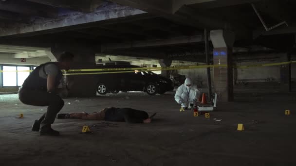 殺人現場を歩き回る男性警察探偵のフルショット 座って 地面に横たわっている死体を見て 手持ちのラジオに話し バックグラウンドで証拠を収集法医学の専門家 — ストック動画
