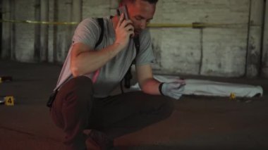 Cinayet mahallinde yere çömelmiş, akıllı telefondan konuşan, kanıtlara bakan, polis arabasının farlarını yakan ve arka planda örtülü kurbanın cesedini gösteren orta boy erkek dedektifin fotoğrafı.