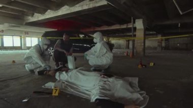 Tanımlanamayan iki adli tıp uzmanının tam yay görüntüsü ve cinayet kurbanının cesedinin yanında çömelmiş ve soruşturmayı tartışan beyaz erkek dedektif.