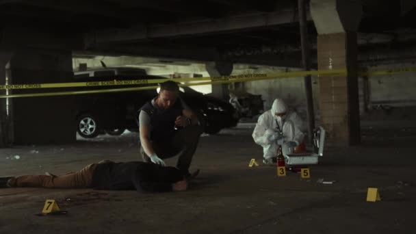 駐車場の犯罪現場で死体を検査し 証拠やサンプルを集めているホワイトオーバーオールの法医学の専門家と話す 防弾ベストの刑事のフルショット — ストック動画