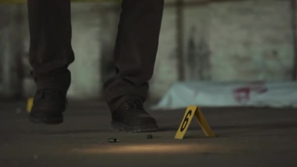凶杀案现场躺在地上的旧弹壳的特写镜头 旁边的黄色法医标记 以及无法辨认的男性侦探的腿 手电筒向他们走来 蹲伏在地上 — 图库视频影像