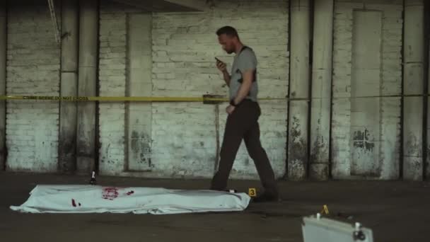 地下ガレージの床に横たわる殺人犠牲者の死体を歩き回り 血液汚れで白いシートで覆われ ポータブルラジオで話す — ストック動画