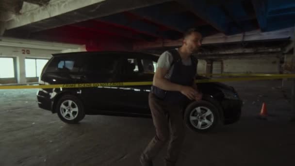犯罪現場の死体に向かって黄色い障壁テープの下の車から歩く防弾ベストの白人男性警察探偵のフルフォローショット トーチを保持し シートを持ち上げて体を見る — ストック動画