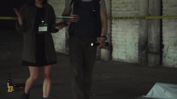 地下駐車場の殺人現場をトーチで歩き Idバッジ付きの女性同僚と話しながら 防弾ベストでコカシアの男性警察探偵の後ろを傾ける中型タイル — ストック動画