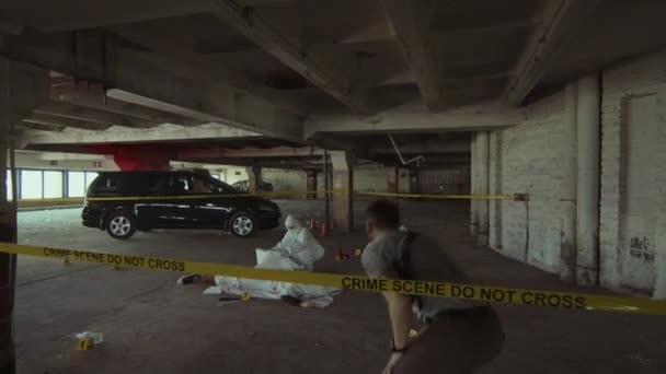 バリアテープの下でハーネスダッキングで銃を持った男性探偵のフルフォローショット 犯罪現場に向かって歩き 死体を見て 死体で作業する白いオーバーオールの法医学の専門家と話す — ストック動画