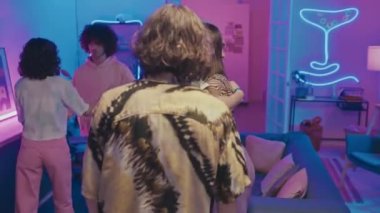 Mavi ve pembe neon ışıklarla ev partisine gelen, birbirini selamlayan ve kucaklayan bir grup sıradan giyinmiş genç kız ve erkek.