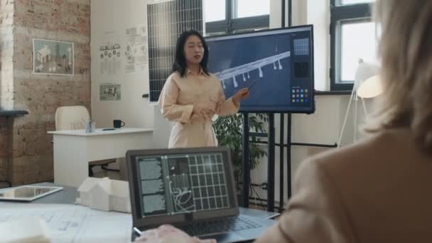 現代のロフトオフィスでマルチメディアスクリーンに革新的なソーラーパネルデザインを提示するアジアの女性エンジニアのミディアムフルオーバーショルダーショット ノートパソコンのリスニングとノッディングを持つ男性同僚 — ストック動画