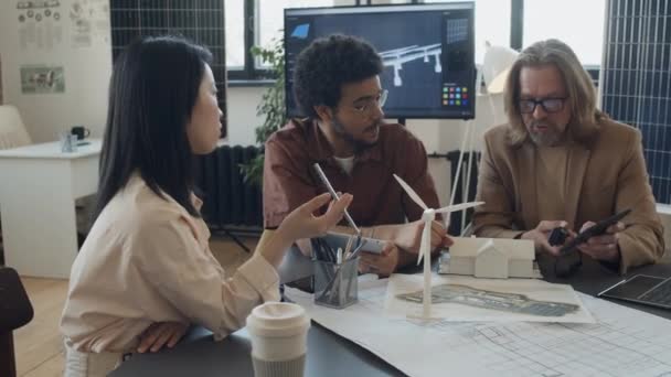 再生可能エネルギーのスタートアップのオフィスのテーブルに座っている3人の多様なエンジニアの中型ショット 風力タービンとの環境に優しい家の設計を議論 背景にソーラーパネル — ストック動画