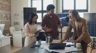 Üç erkek ve kadın mühendisin planlarla ve rüzgar türbinli küçük ölçekli ev modeliyle masanın etrafında durup tasarımıyla ilgili tartışırken orta ölçekli tam kavisli bir fotoğraf.