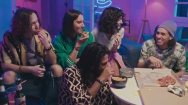 Ortalama olarak beş etnik kız ve erkek kanepede oturmuş, pizza yiyor, patates cipsi yiyor, bira içiyor, TV 'de komedi programı izliyor, sohbet ediyor ve parmaklarını gösteriyorlar.