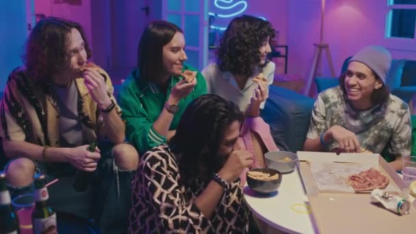 五个多民族女孩和男人坐在房间里的沙发上 吃披萨 喝啤酒 看电视上的喜剧节目 聊天和指指点点 拍到一半的全景 — 图库视频影像