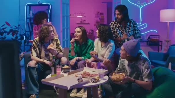 在霓虹灯照亮的房间里 穿着休闲装坐在沙发上 喝啤酒 吃外卖意大利香肠披萨 笑着聊天的年轻男女朋友们的全景照片 — 图库视频影像