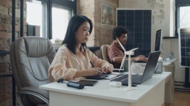 Yenilenebilir enerji şirketinin çatı katında masa başında oturan Asyalı kadın mühendisin orta kavisli görüntüsü ve arka planda rüzgar türbini ve güneş paneli olan grafik tabletine yeni ürün konsepti çizmesi