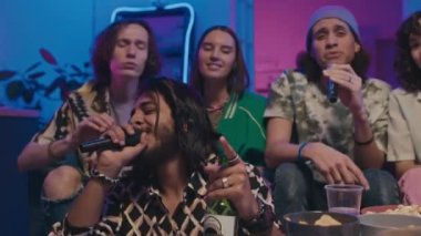 Orta boy iki Orta Doğulu gencin bira şişeleriyle kanepede oturup mikrofonlara karaoke şarkıları söylerken ve arkadaşlarının müziğe gülüp sallanırken.