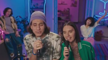 Orta boy Orta Doğulu bir adam ve Kafkasyalı bir kızın ev partisinde mikrofonlara sarılıp karaoke şarkıları söylemesi ve dans etmesi ve arkadaşlarının arka planda gülüp sallanmaları.