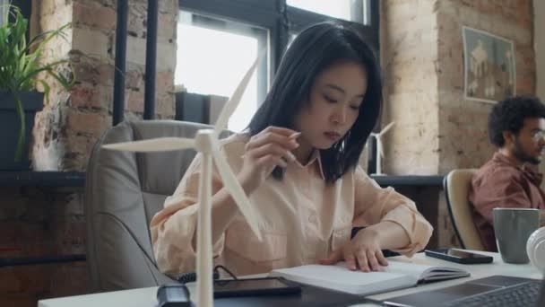 中国女性产品设计师坐在阁楼办公室的桌子前 看着手提式太阳能充电器 然后在笔记本上写字 在电脑上打字 还有展望中的风力涡轮机模型 — 图库视频影像