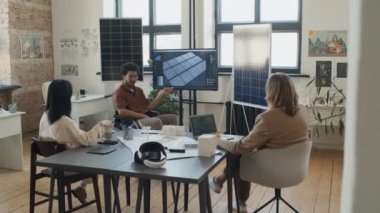 Yenilenebilir enerji şirketinin ofisinde masanın etrafında oturan, multimedya ekranına bakan ve güneş enerjisi projesini tartışan etnik çeşitliliğe sahip erkek ve kadın mühendislerin tam karesi.