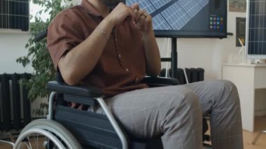 Tekerlekli sandalyedeki genç melez erkek mühendisin yenilenebilir enerji şirketinde yaptığı toplantıda konuşurken, ekranda güneş enerjisi projesini meslektaşlarına sunarken orta eğik bir görüntü.