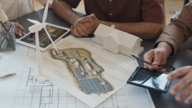 Mimari planlar, küçük ölçekli ev, rüzgar türbini, güneş pili, diyagramı olan tablet ve karbon nötr projesini tartışan çeşitli mühendislerin el yapımı resimleri.