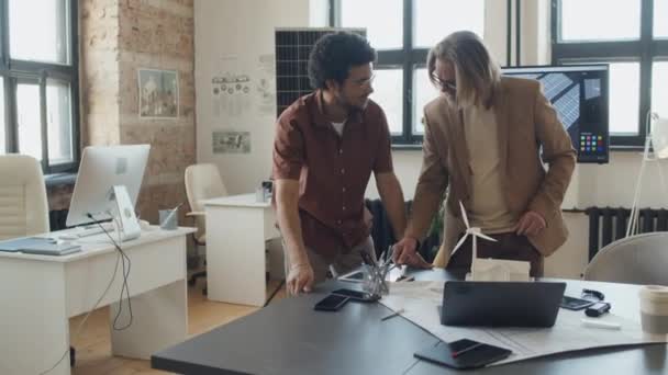 オフィスのテーブルに立っている2人の多様な男性建築家の中型ショット 風力タービンでゼロカーボン住宅プロジェクトと中国の同僚がコンピュータで机に向かって歩くことについて話し合う — ストック動画
