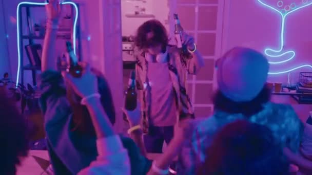 中镜头的年轻Dj在学生宿舍聚会上演奏 带着迪斯科舞会和霓虹灯 带着一瓶啤酒跳舞 还有一群年轻人跟在后面挥手 — 图库视频影像