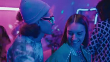 Melez, kıvırcık saçlı, bereli, gözlüklü ve beyaz bir kızın ev partisinde mavi ve pembe neon ışıklı ve disko topuyla dans ettiği orta boy yakın plan bir fotoğraf.