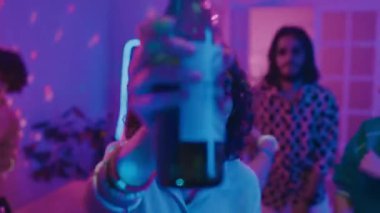 Kıvırcık saçlı, kalça güneş gözlüklü, neon kolyeli, elinde bira şişesi olan, partide dans eden, kameraya gülümseyen, arkadaşlar ve DJ 'in arka planda eğlendiği orta boy yakın çekim.