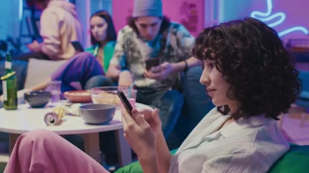 在家庭派对上 带着智能手机坐在豆袋椅子上 在社交网络上翻阅新闻资料 背景朋友的中等选择性聚焦侧影 — 图库视频影像