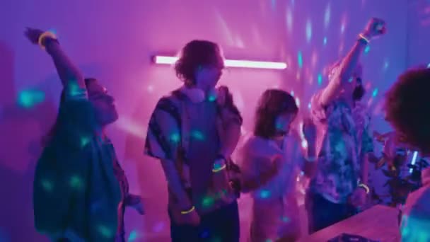 穿着便装的年轻人在家庭聚会上玩得很开心 拿着啤酒瓶和Dj转盘围着桌子跳舞和跳跃 开着蓝色和粉色的霓虹灯迪斯科舞厅 — 图库视频影像