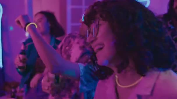 エネルギッシュに踊り ハウスパーティーで ブルーとピンクのネオンライトで一緒に楽しんで ビールボトルを持って楽しいメガネを着ている若い女の子と男の子のミディアムクローズアップショット — ストック動画