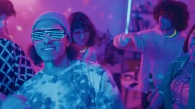 Öğrenci partisinde neon gözlüklü genç erkek öğrencinin enerjik bir şekilde dans edişinin, çeşitli arkadaşlarının birayla dans edişinin, DJ 'in pembe ve mavi disko topu ışıklarıyla dans edişinin orta boy yakın çekimi.