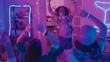 Genç DJ 'in pikaplarda popüler bir şarkı çalarken, misafirlerle enerjik bir şekilde zıplarken ve müzik ritmine el sallarken.