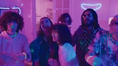 Parlak elbiseli genç erkek ve kadın hippilerden oluşan orta ölçekli bir grup ve ev partisinde enerjik bir şekilde dans eden, kameraya bakan ve arka planda DJ çalan bir grup.