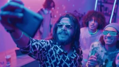 Ortadoğulu genç bir adamın güneş gözlüklü ve çeşitli arkadaşları bira şişeleriyle dans ederken ve akıllı telefondan grup selfie çekerken, DJ arka planda neon ışığı eşliğinde çalarken.